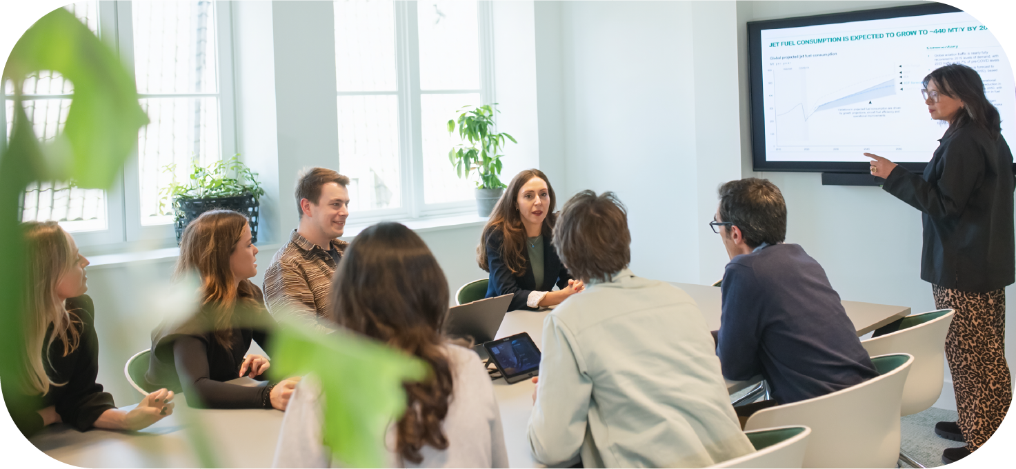 A group of professionals engaged in a meeting around a conference table, with a presenter pointing towards a screen displaying a graph about jet fuel consumption projections.